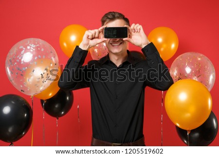 Smiling young man in black classic shirt covering eyes with mobile phone with blank black empty screen on red background air balloons. Women's Day Happy New Year birthday mockup holiday party concept