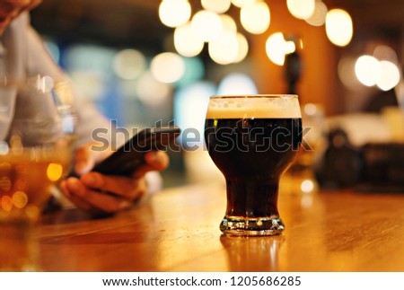 a glass of draft or craft beer in a mug, focus at the back glass of picture, black beer with smooth foam, bokeh background in night club,a hand of people play smartphone, with copy space.