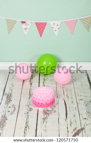 Beautifully designed pink ombre butter icing birthday party cake on distressed wooden white washed floor and green background with green and pick flag bunting and balloons