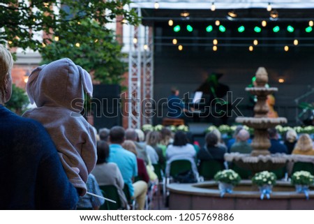 Mom is holding her son or daughter in hoodie with bunny ears and enjoying the culture. Romantic evening in concert venue with jazz musicians playing songs on stage. Beautiful wedding celebration
