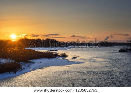 Beautiful russian early winter landscape with non frozen river, snow-covered riverside and dried grass at sunset. Scenic nature background