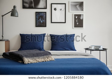 Close-up of bed with blue bedding and dark colored blanket. Bedside table with books and coffee next to it. Gallery of framed art on the wall. Real photo concept