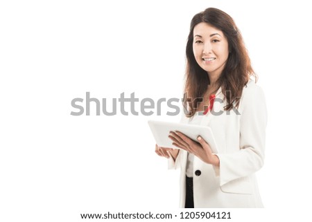 smiling asian businesswoman with aids awareness red ribbon on jacket using tablet and looking at camera isolated on white