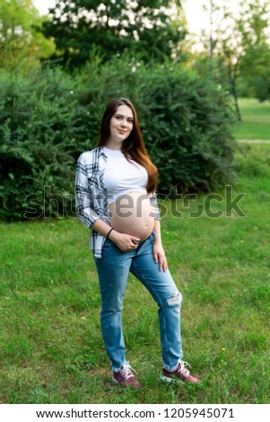 Beautiful smiling and stylish pregnant woman in jeans and checked shirt holding hands on tummy. Brunette mom touching, embracing stomach posing in park. Mother spending time outdoors.