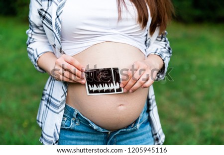 Crop of stylish pregnant woman in jeans and checked shirt holding first photo screen of baby. Brunette mom touching, embracing stomach posing in park. Mother spending time outdoors.