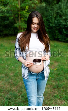 Portrait of stylish pregnant woman in jeans and checked shirt holding first photo screen of baby. Brunette mom touching, embracing stomach posing in park. Mother spending time outdoors.