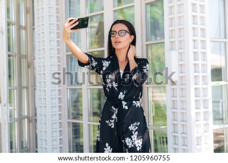Close-up of a beautiful young girl wearing glasses has a European appearance, dark straight hair, wearing a silk outfit. A woman makes selfie on her smartphone, smiling.