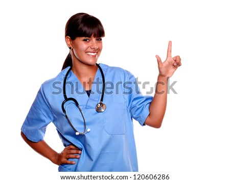 Portrait of a smiling young nurse on blue uniform with a stethoscope looking at you while is pointing up standing over white background - copyspace
