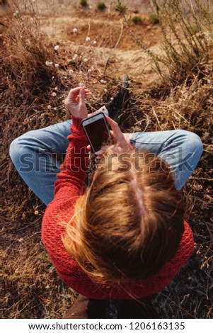 A young ginger girl photographer in a red sweater taking pictures of flowers on a rocky coast.