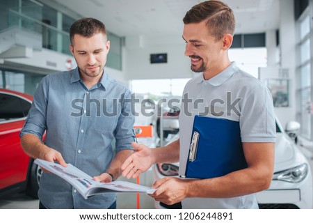 Picture of two men standing inside. They hold journal and look at it. People smile. They stand at red car. Man in white shirt points on journal and smile.