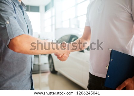 Picture of two young men shaking hands in front of white car. They made an agreement. Customer and seller works together. Cut view.