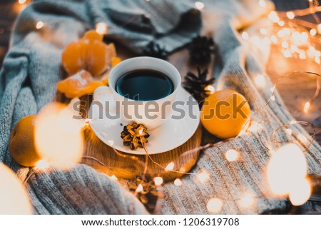 A white cup with a saucer in which hot coffee is poured. The cup is on a wooden background, around which lies a gray knitted textile. Also in the photo there are tangerines, pine cones and a garland .