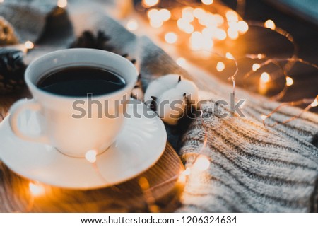 A white cup with a saucer in which hot coffee is poured. The cup is on a wooden background, around which is a gray knitted textile. Also in the photo there are tangerines, cotton flowers and a garland