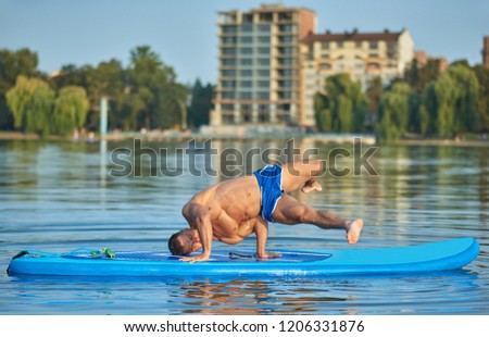 Handsome young boy doing hand standing position on paddle board during hot summer day. Sportsman with perfect figure, sporty body, wearing blue shorts, training outside. Concept of healthy lifestyle.