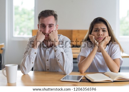 Bored male and female students studying in classroom. Young man and woman sitting at table, leaning heads on hands and looking at camera. Education concept. Front view.