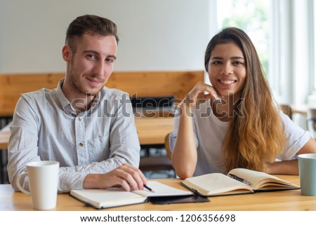 Positive students studying and posing at camera. Young man and woman using notebooks and sitting at desk in classroom or library. Education and friendship concept. Front view.