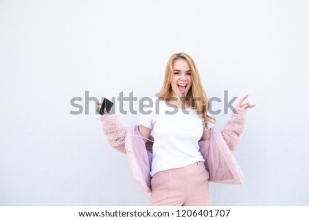 Emotional happy girl dressed in a pink coat and a white T-shirt, listens to music in the headphones with a smartphone in her hands, looks camera and smiles.Sweet emotional blonde on a white background