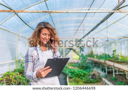 View of a Young attractive woman working at the plants nursery using smartphone. Picture of happy young woman standing in greenhouse talking by phone. Looking aside.