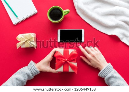 Female hands, a woman holding a gift and uses smart phone. Red table, white smart phone, gifts boxes for the holidays, background with copy space for advertisement, top view
