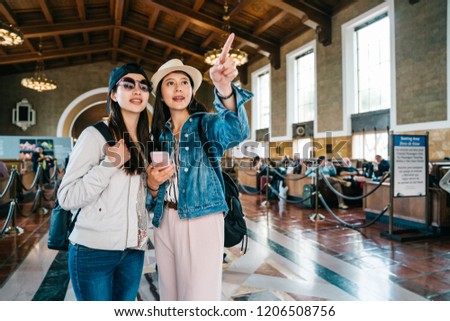 joyful female travelers pointing to the sign and discussing the right way in union station. friendship concept of young girls. Young ladies using smartphone app visiting in US.