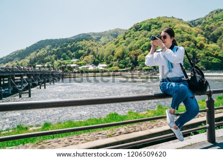 elegant photographer sitting on the handrail and taking picture of the Togetsukyo bridge. traveling lens man hobby love taking scenery photo by slr camera. green mountains on a sunny day in Japan.
