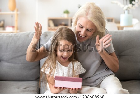 Smiling grandmother making surprise to cute little granddaughter giving gift box, excited small girl get birthday present from granny, happy older and younger generation celebrate at home together
