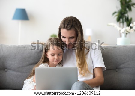 Mother and daughter sit on couch using laptop watching video or cartoon online, cute little girl spend time with mom, relax at home browsing computer or shopping together. Young and technology concept