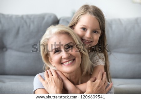 Portrait of cute little granddaughter piggyback smiling grandmother posing for family picture, granny and grandchild hugging having fun together, small girl embrace loving granny spending time at home