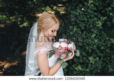 Beautiful and gentle bride stands with a bouquet in nature with green trees. Wedding portrait. Wedding photography.