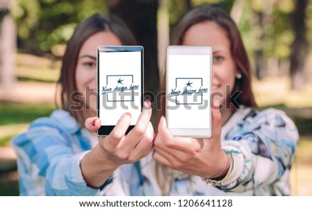 Women friends showing smartphones with their selfie photos taked over a forest background. Customizable mobile screens