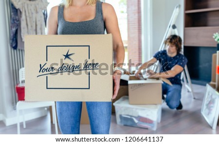 Woman carrying moving box while her husband unpacks. Customizable box design Royalty-Free Stock Photo #1206641137