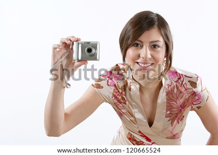 Attractive young woman using a digital photographic camera while standing isolated against a plain white background, smiling at the camera.