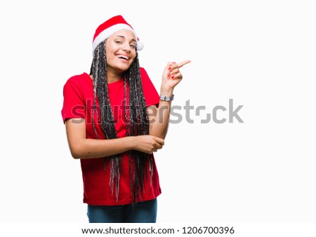 Young braided hair african american girl wearing christmas hat over isolated background with a big smile on face, pointing with hand and finger to the side looking at the camera.
