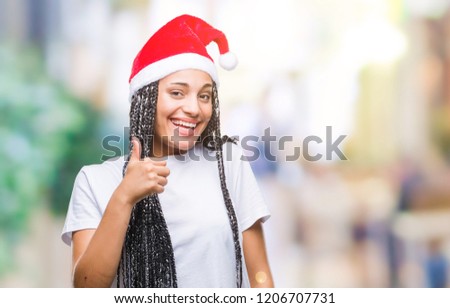 Young braided hair african american girl wearing christmas hat over isolated background doing happy thumbs up gesture with hand. Approving expression looking at the camera with showing success.