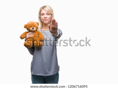Young beautiful blonde woman holding teddy bear plush over isolated background with open hand doing stop sign with serious and confident expression, defense gesture