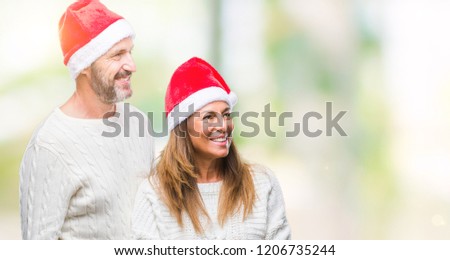 Middle age hispanic couple wearing christmas hat over isolated background looking away to side with smile on face, natural expression. Laughing confident.