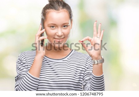 Young beautiful caucasian woman talking on smartphone over isolated background doing ok sign with fingers, excellent symbol