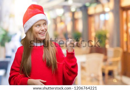 Young beautiful girl wearing christmas hat over isolated background smiling with happy face looking and pointing to the side with thumb up.