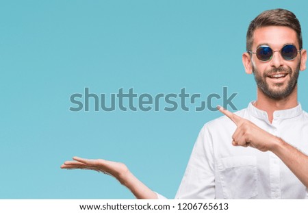Young handsome man wearing glasses over isolated background amazed and smiling to the camera while presenting with hand and pointing with finger.
