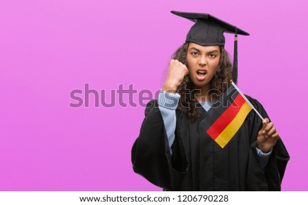 Young hispanic woman wearing graduation uniform holding flag of Germany annoyed and frustrated shouting with anger, crazy and yelling with raised hand, anger concept