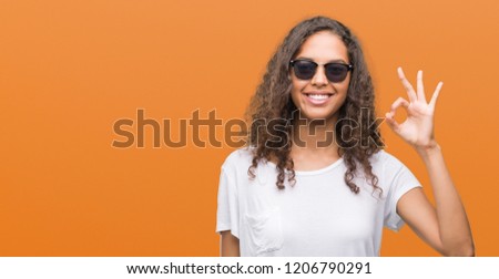 Young hispanic woman wearing sunglasses smiling positive doing ok sign with hand and fingers. Successful expression.