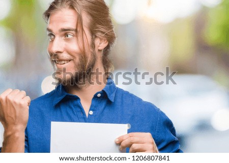Young handsome man with long hair over isolated background holding blank paper pointing and showing with thumb up to the side with happy face smiling