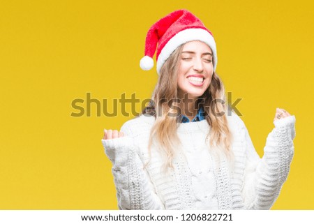 Beautiful young blonde woman wearing christmas hat over isolated background very happy and excited doing winner gesture with arms raised, smiling and screaming for success. Celebration concept.