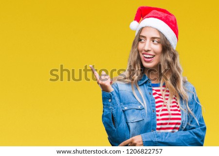 Beautiful young blonde woman wearing christmas hat over isolated background with a big smile on face, pointing with hand and finger to the side looking at the camera.