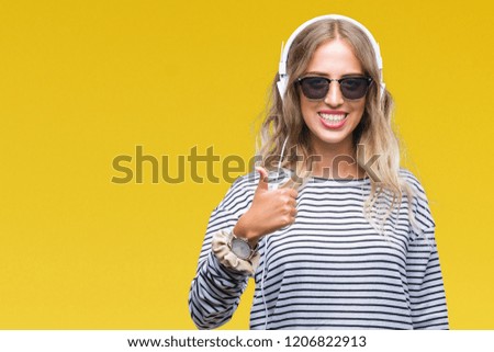 Beautiful young blonde woman wearing headphones and sunglasses over isolated background doing happy thumbs up gesture with hand. Approving expression looking at the camera with showing success.