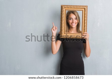 Beautiful young woman over grunge grey wall holding vintage frame surprised with an idea or question pointing finger with happy face, number one