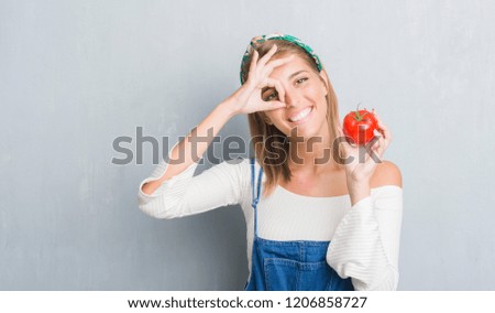 Beautiful young woman over grunge grey wall eating fresh tomato with happy face smiling doing ok sign with hand on eye looking through fingers