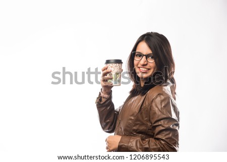 Pretty Indian young girl having coffee while wearing spectacles and speaking over phone, standing isolated over white background