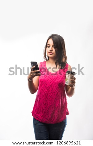 Pretty Indian young girl having coffee while wearing spectacles and speaking over phone, standing isolated over white background
