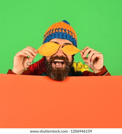 Hipster with beard and smile closes eyes with leaf. Autumn and cold weather concept. October and November time idea. Man holds yellow cherry tree leaves on green and orange background, copy space